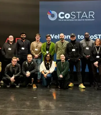 Group of about 18 people posing for a photo on a stage. They are standing and kneeling in front of a blue backdrop with the CoSTAR logo and the text “Welcome to CoSTAR Evolve!” Many wear lanyards and casual to smart-casual clothing