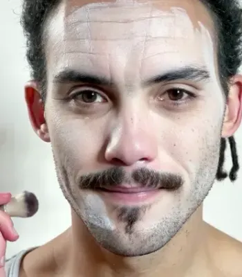 A man applying makeup with a brush, white powder on his forehead and cheeks, sporting a mustache and goatee, wearing a light gray sleeveless shirt.