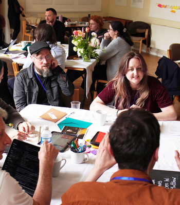 a group of about 20 diverse people in a room sat round tables taking part in a workshop with animated expressions