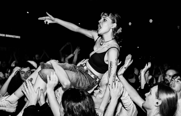 Black-and-white photo of a female crowd-surfing performer being carried above a packed concert crowd. She wears a black crop top and jeans, has tattoos on her left arm, and points outward with her right arm while fans reach up to support her