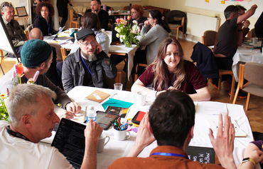 a group of about 20 diverse people in a room sat round tables taking part in a workshop with animated expressions