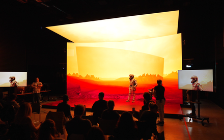 The CoSTAR Screen Lab LED stage is lit up displaying a Martian landscape. An actor in a spacesuit is on the stage. A cameraman is shooting the scene from the side. A group of spectators watch from seats in front of the stage.