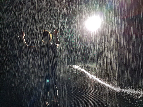 Photo of person holding hands up as if dancing in the rain with what looks like rain and a bright spotlight behind them 