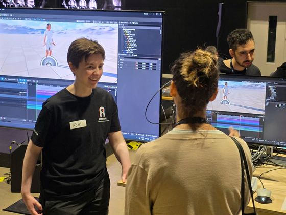 Two attendees stand in front of large monitors displaying animation software and a stylised 3D character environment during the AI for Creativity workshop at CoSTAR Screen Lab, Studio Ulster.
