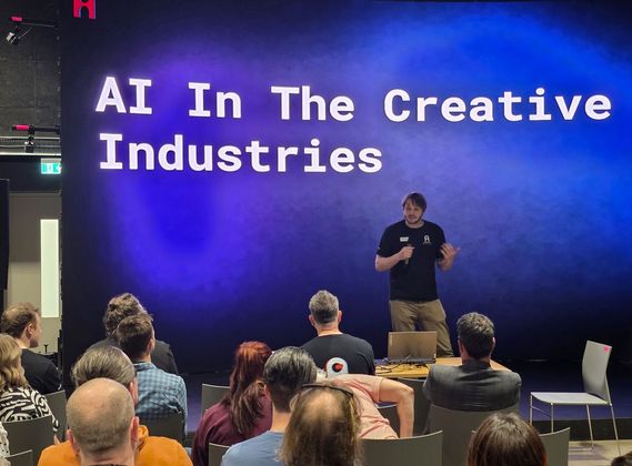 A speaker presents to a seated audience on stage beneath a large screen displaying the words “AI In The Creative Industries” during a workshop at CoSTAR Screen Lab, Studio Ulster.