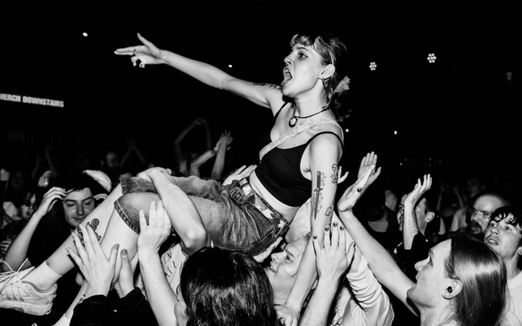 Black-and-white photo of a female crowd-surfing performer being carried above a packed concert crowd. She wears a black crop top and jeans, has tattoos on her left arm, and points outward with her right arm while fans reach up to support her
