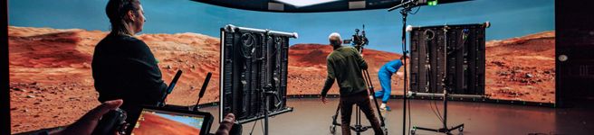 A film crew works inside a virtual production studio, filming in front of a curved LED wall displaying a desert-like Mars landscape beneath a large overhead soft light.