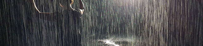 Photo of person holding hands up as if dancing in the rain with what looks like rain and a bright spotlight behind them 