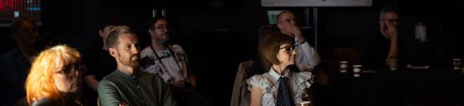 a group of people sitting at tables in a darkened room focusing on something to the right of screen two men and two women are in view
