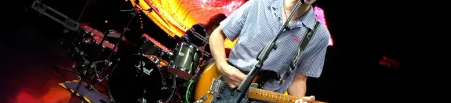 Guitarist singing into a microphone while playing an electric guitar on stage, with a drummer and colorful background lights.