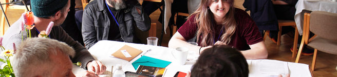 a group of about 20 diverse people in a room sat round tables taking part in a workshop with animated expressions