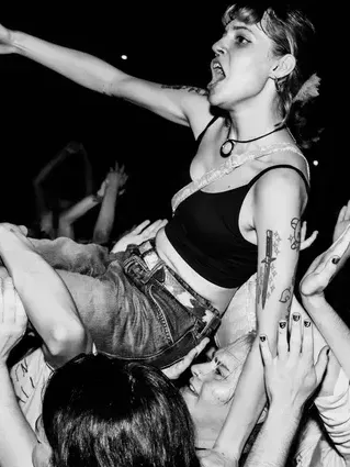 Black-and-white photo of a woman crowd-surfing at a concert, standing above a sea of raised hands. She wears a black crop top, jeans, and a choker, with fans surrounding and supporting her.