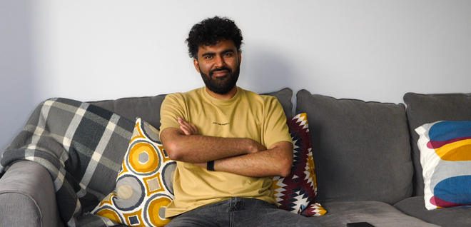 A man with dark curly hair and a beard sits on a gray couch in a living room, arms crossed, wearing a tan T-shirt. He is flanked by colorful patterned throw pillows and a plaid blanket draped over the arm of the sofa.