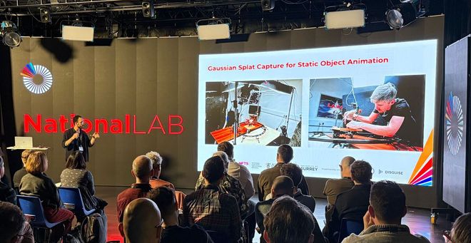A view of an audience from the back in front of a large VP screen with a presentation showing a man placing a puppet snail in front of a small camera