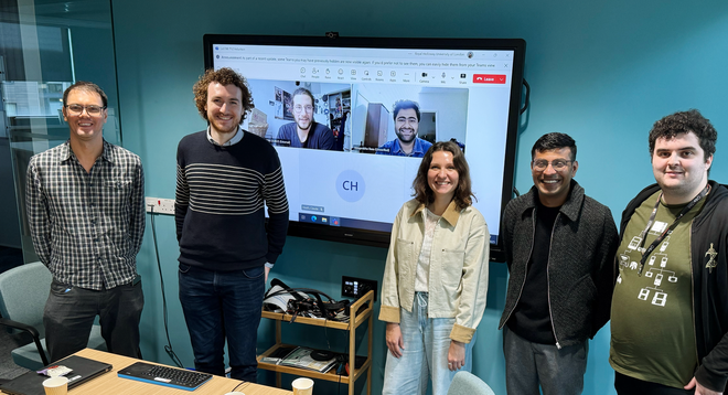Five CoSTAR PhD students stand in a teal-walled room, smiling at the camera. They pose in front of a large wall-mounted touchscreen showing a multi-person video conference. A small rolling cart with cables and a keyboard sits beneath the screen, and a desk with a cup and papers is in the foreground.