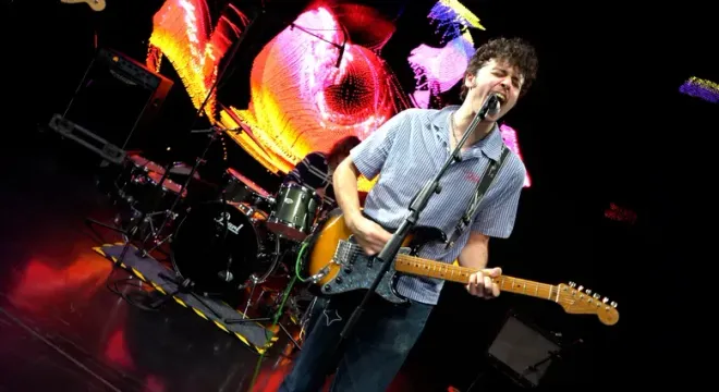 Guitarist singing into a microphone while playing an electric guitar on stage, with a drummer and colorful background lights.