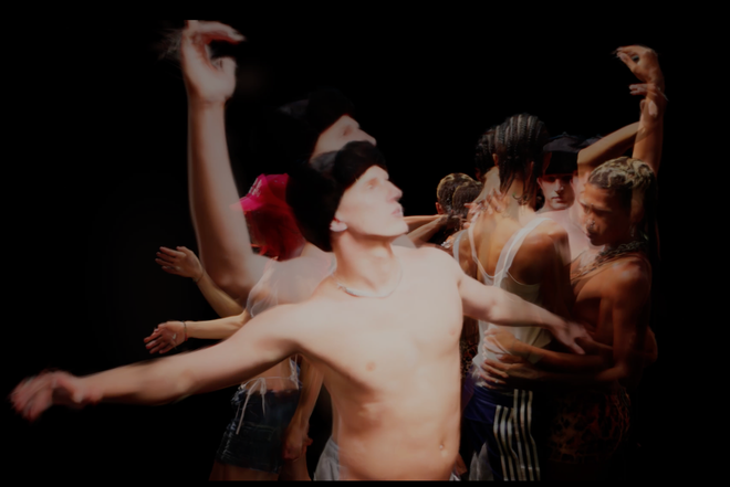 Dynamic contemporary dance on a dark stage: a shirtless male dancer with arms extended in the foreground, surrounded by a blurred group of dancers in varied outfits.