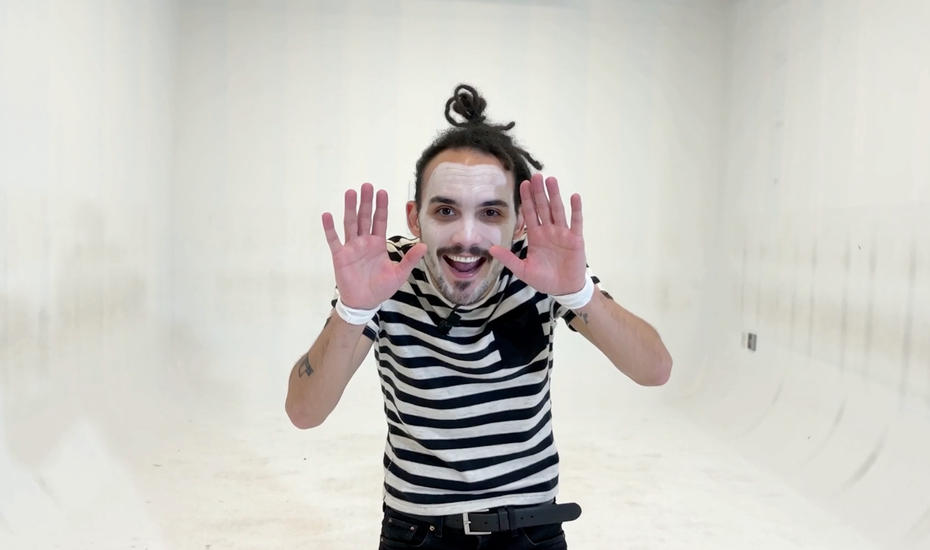 photo of a man with dark skin dressed as a mime smiling to camera and miming with his palms up and facing camera