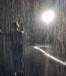 Photo of person holding hands up as if dancing in the rain with what looks like rain and a bright spotlight behind them