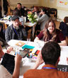 a group of about 20 diverse people in a room sat round tables taking part in a workshop with animated expressions