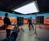 A film crew works inside a virtual production studio, filming in front of a curved LED wall displaying a desert-like Mars landscape beneath a large overhead soft light.