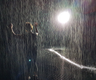 Photo of person holding hands up as if dancing in the rain with what looks like rain and a bright spotlight behind them 