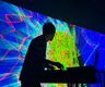 an image showing a silhouette of a man using a laptop and mixing desk with a colourful computer generated graphic showing on large screen as a backdrop